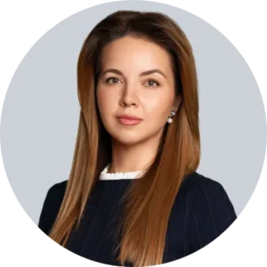 Professional headshot of a woman with long brown hair, wearing a dark blazer and white blouse, against a neutral gray background.