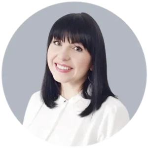 Smiling woman with shoulder-length black hair in a white blouse, head-and-shoulders portrait against a light gray background.