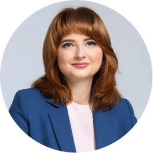 Portrait of a smiling woman with auburn hair wearing a blue blazer and light pink top, against a light gray background.