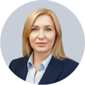 Professional portrait of a blonde woman in a navy blazer and light blue shirt, facing the camera with a neutral expression, against a light blue-gray circular background.